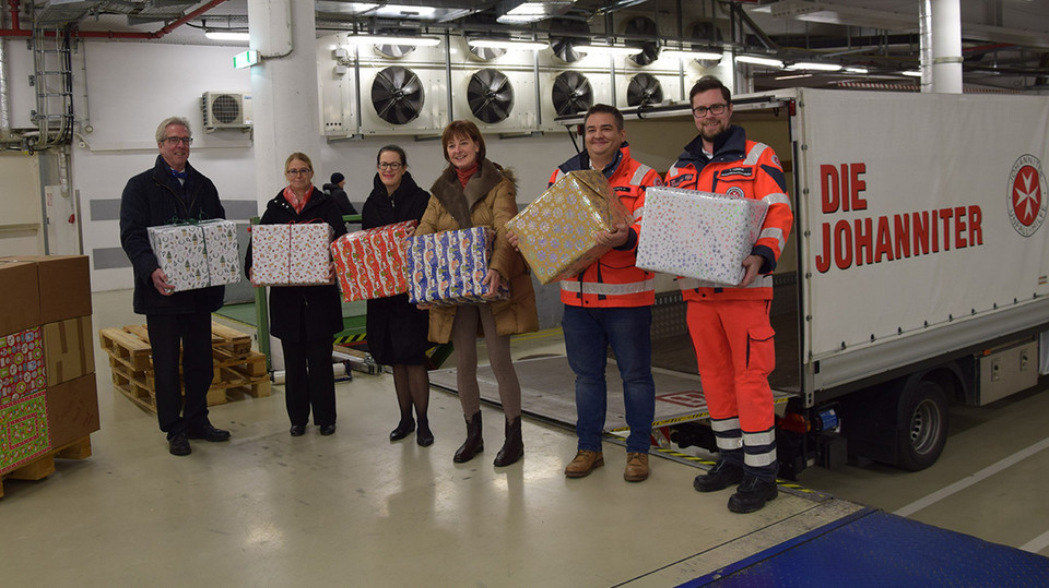Sechs Personen halten Weihnachtsgeschenke in der Hand und stehen vor einem kleinen LKW