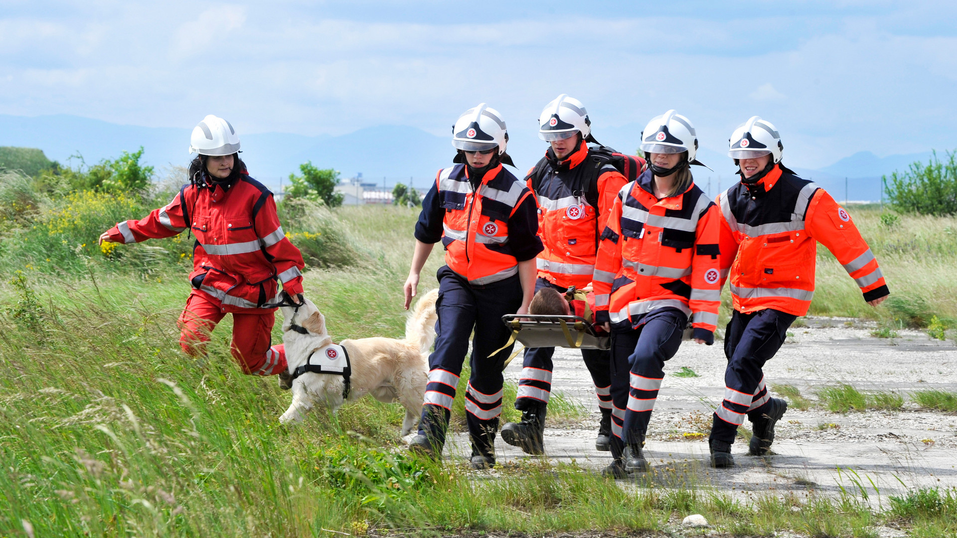 Vier Johanniter tragen eine verletzte Perosn auf einer Trage. Daneben ist eine Johanniterin mit ihrem Rettungshund.