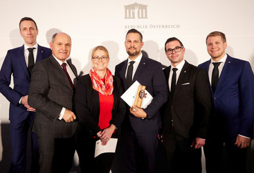 Fünf Männer in dunklen Anzügen und in der Mitte eine Frau stehen vor einer weißen Wand, der Mann in der Mitte hält eine hölzerne Trophäe und eine Urkunde in der Hand 