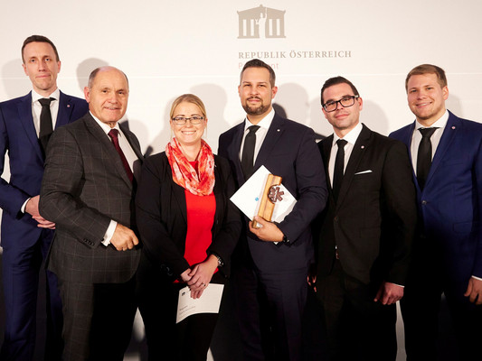 Fünf Männer in dunklen Anzügen und in der Mitte eine Frau stehen vor einer weißen Wand, der Mann in der Mitte hält eine hölzerne Trophäe und eine Urkunde in der Hand 