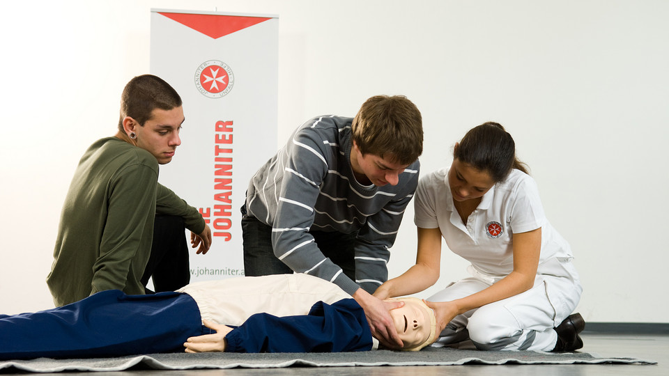 Eine Kurslehrerin zeigt zwei Jugendlichen die richtige Lagerung an einer Erste-Hilfe-Puppe.