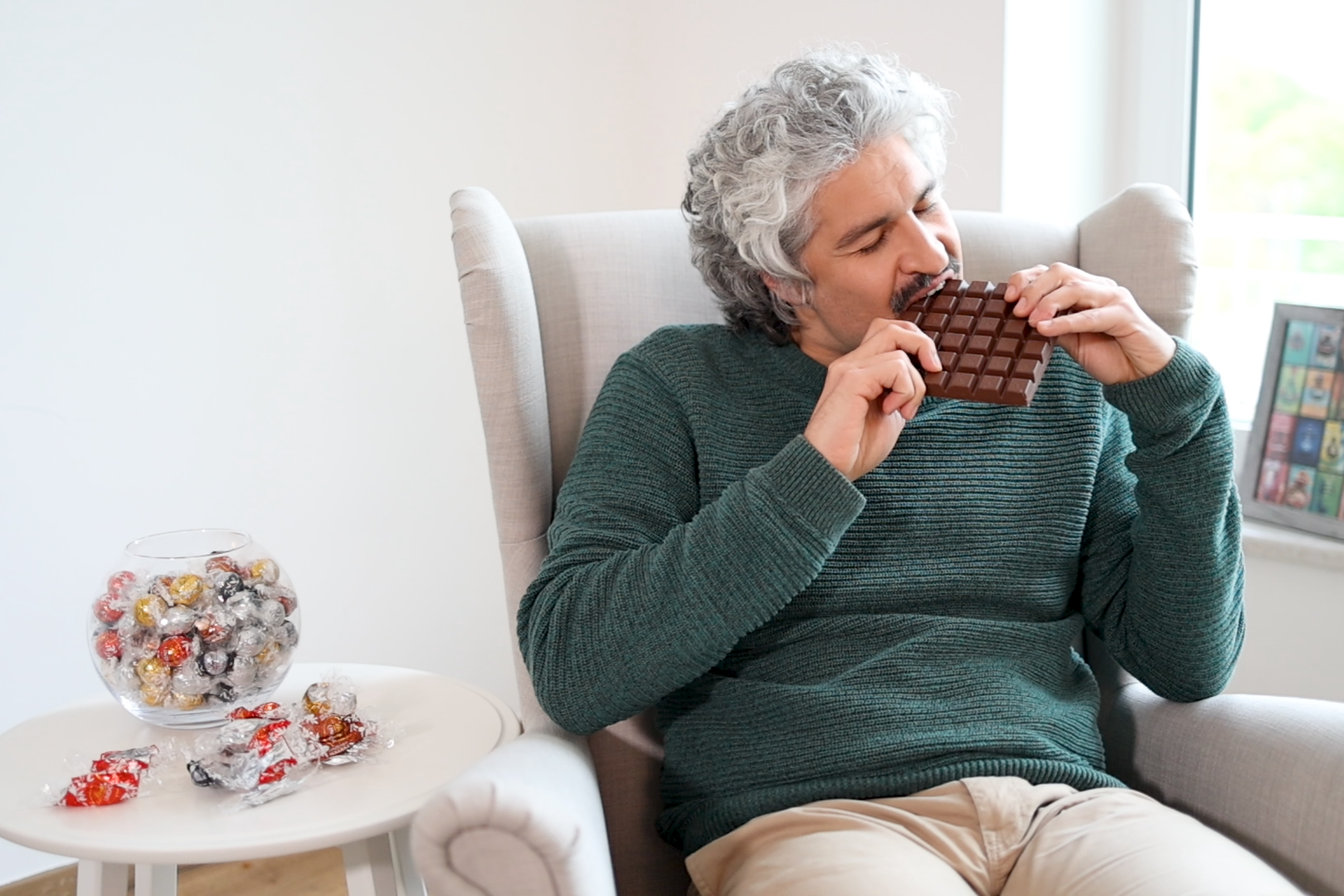 Ein Mann, Aleksandar, sitzt in einem Ohrensessel beißt in eine Tafel Schokolade. Neben ihm steht ein Glas mit einzeln verpackten Pralinen.