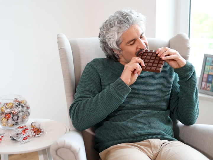 Ein Mann, Aleksandar, sitzt in einem Ohrensessel beißt in eine Tafel Schokolade. Neben ihm steht ein Glas mit einzeln verpackten Pralinen.