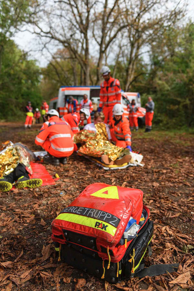 Ein Rettungsrucksack liegt offen im Vordergrund am Waldboden, während im Hintergrund Patient:innen von Sanitäter:innen versorgt werden.
