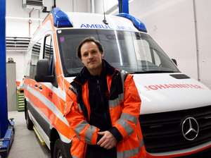 Mann in Uniform steht vor Rettungsauto, im Hintergrund ist eine Autowerkstätte zu sehen. 