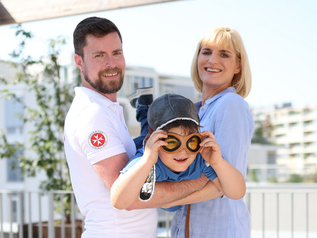 Ein Kind mit einer Fliegerhaube wird von einer Frau und einem Mann getragen als ob er fliegen würde.