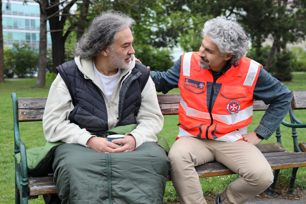 Ein Johanniter spricht mit einem Wohnungslosen, der mit einem Schlafsack auf der Parkbank sitzt.
