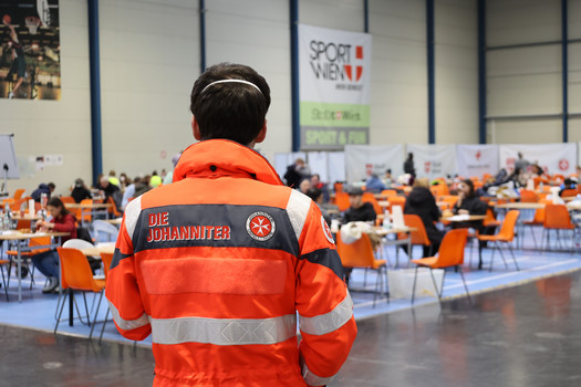 Ein Johanniter schaut auf die aufgestellten Tische in der Sport & Fun Halle, wo ankommende Menschen zur Ruhe kommen.