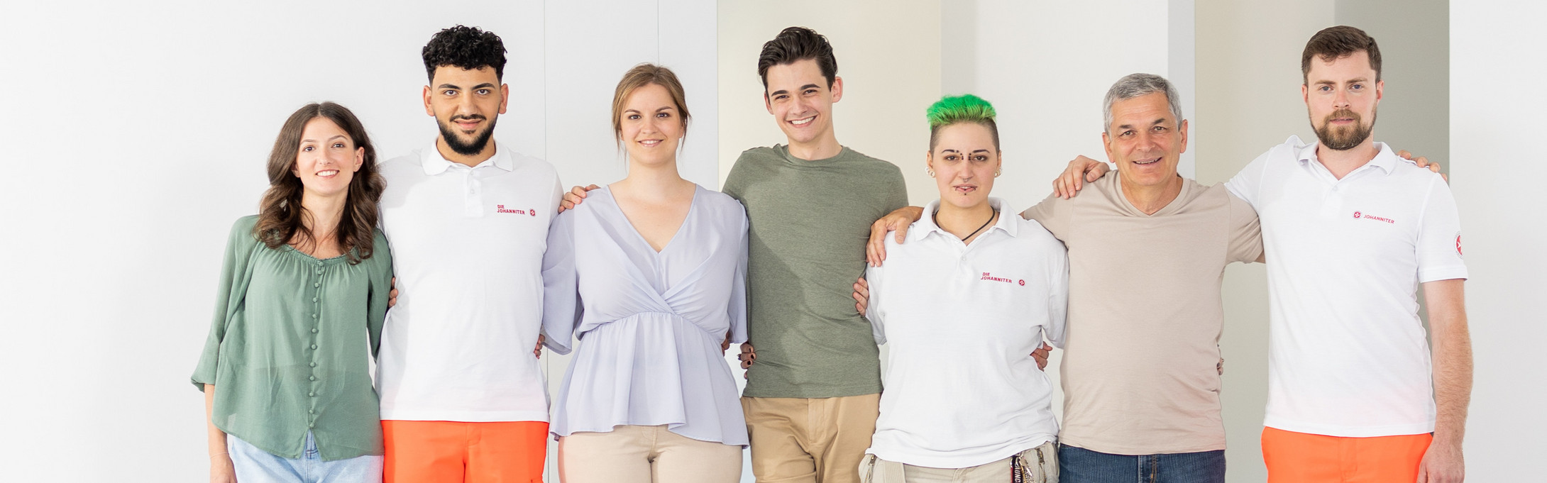 Eine Gruppe von sieben Menschen - jung bis alt, Frauen und Männer - gehen Arm in Arm lächeln auf die Kamera zu. Darunter befinden sich auch zwei Johanniter.