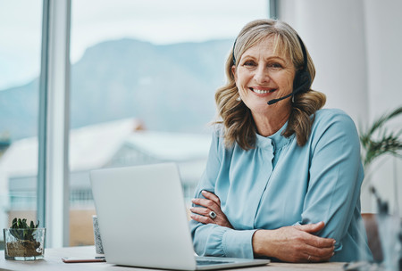 Eine ältere Frau sitzt vor dem Laptop, hat ein Headset auf und lächelt in die Kamera.