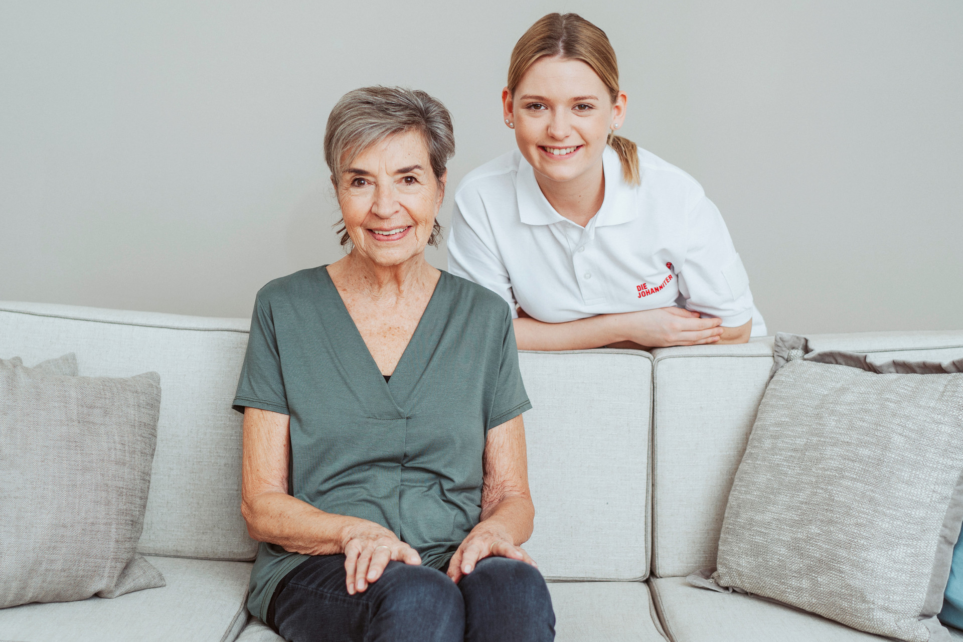 Eine Patientin sitzt auf einem Sofa. Eine Pflegerin stützt sich hinter ihr mit den Unterarmen an der Rückenlehne ab und schaut ihr über die Schulter.