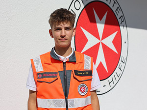 junger Sanitäter in Uniform steht vor einer weißen Wand mit großem Runden Johanniter-Emblem 