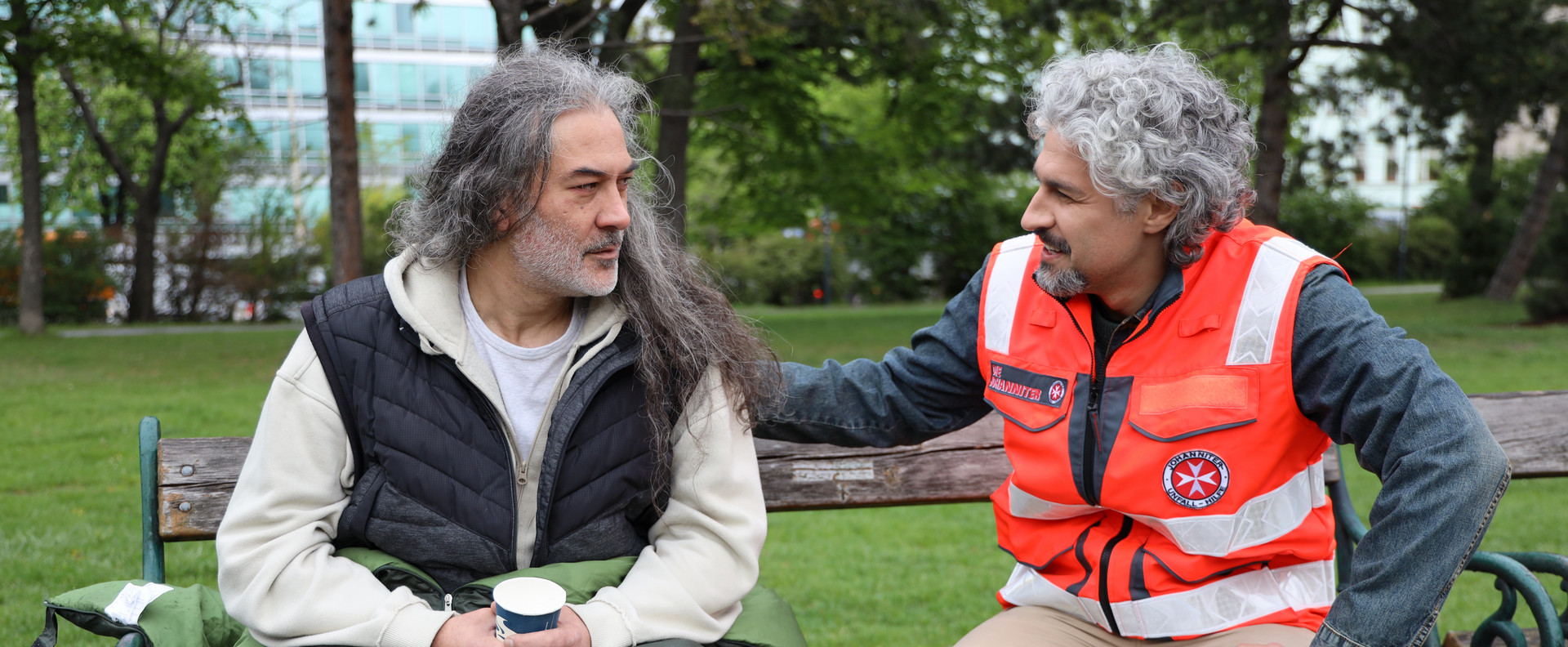 Ein Johanniter spricht mit einem Wohnungslosen, der mit einem Schlafsack auf der Parkbank sitzt.