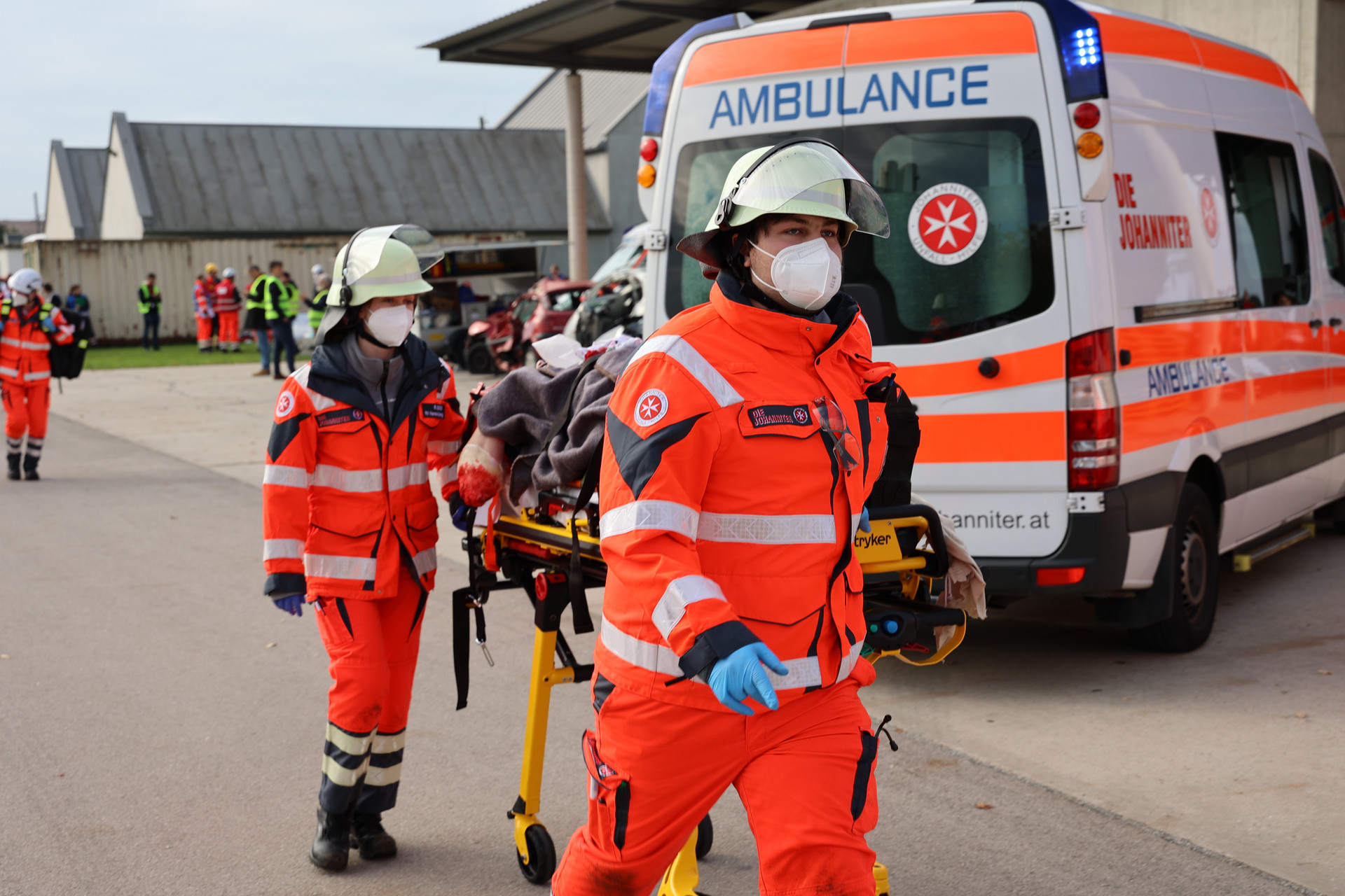 Zwei Johanniter transportieren eine schwerverletzte Person auf einer Fahrtrage ab. Im Hintergrund steht ein Krankenwagen.