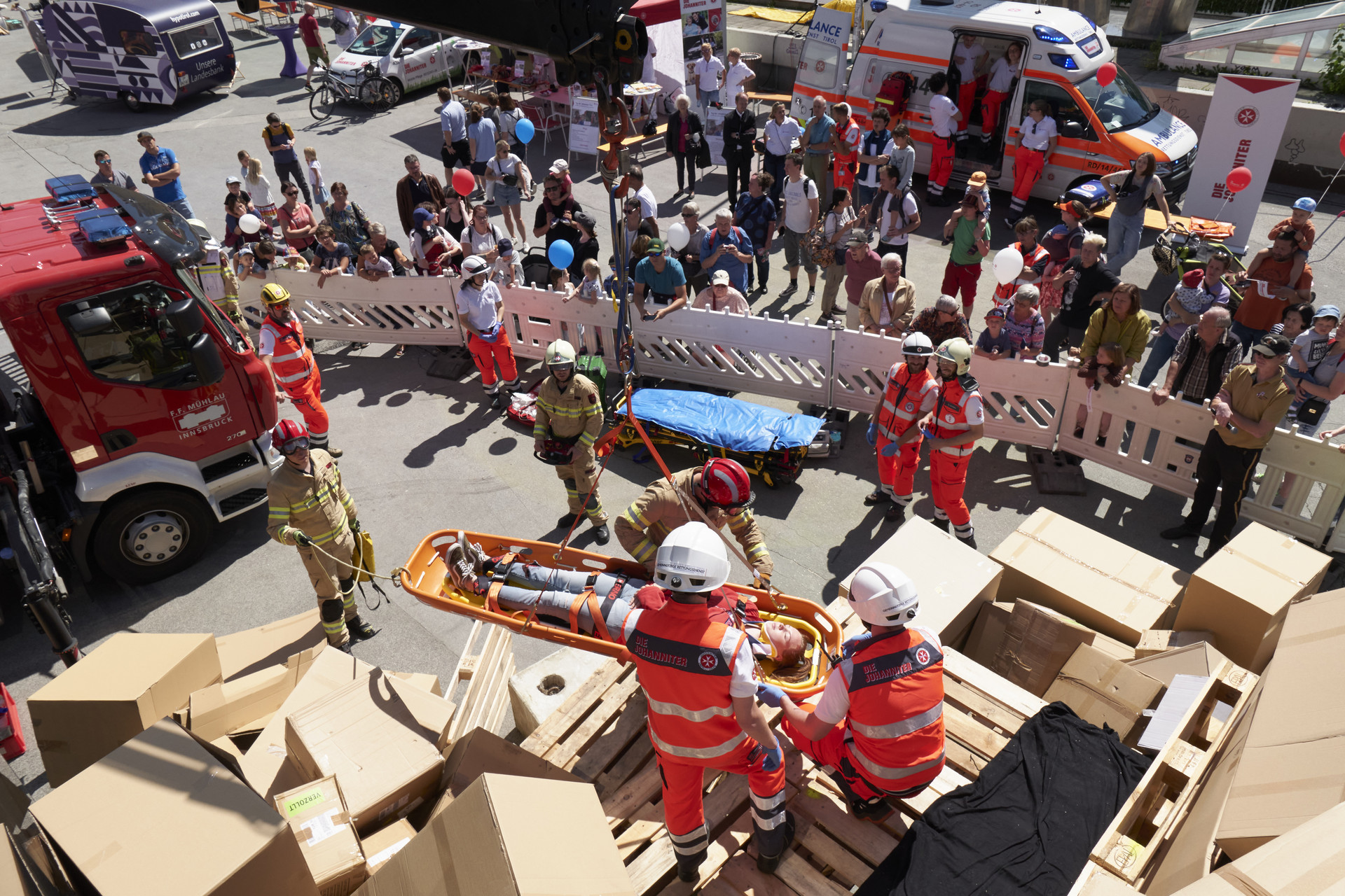 Rettungskräfte bergen gemeinsam mit Feuerwehrleuten die verletzte Person aus dem Berg aus Kartons mit einem Kran. Hinter den Absperrungen stehen die Zuschauer:innen der Übung.