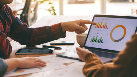 Drei Personen sitzen in einem Meeting vor einem Laptop. Eine Person zeigt auf eine Statistik, die am Laptop gezeigt wird.