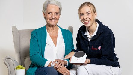 Eine Klientin mit dem Hausnotruf-Armband sitzt neben einer Johanniterin, die die Basisstation in der Hand hält.