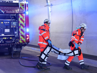 zwei Sanitäter tragen eine Übungspuppe, im Hintergrund sieht man ein Feuerwehrauto mit Blaulicht