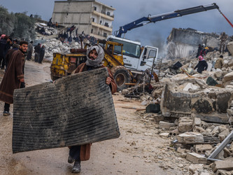 Mann trägt eine schwarze Matraze, im Hintergrund sind zerstörrte Gebäude zu sehen