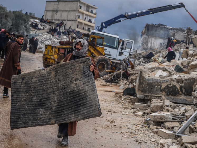 Mann trägt eine schwarze Matraze, im Hintergrund sind zerstörrte Gebäude zu sehen