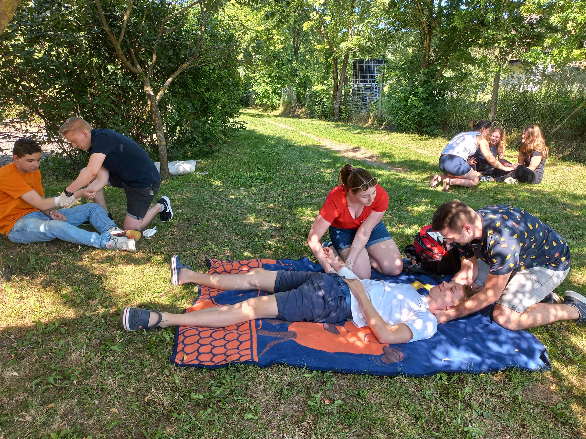 mehrere Personen sitzen in Gruppen beisamen auf einer Wiese, gemeinsam üben sie Erste-Hilfe-Techniken