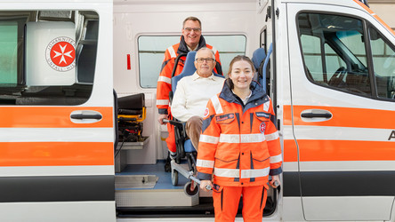 Zwei Johanniter heben einen Patienten auf einem Tragsessel aus dem Krankenwagen.