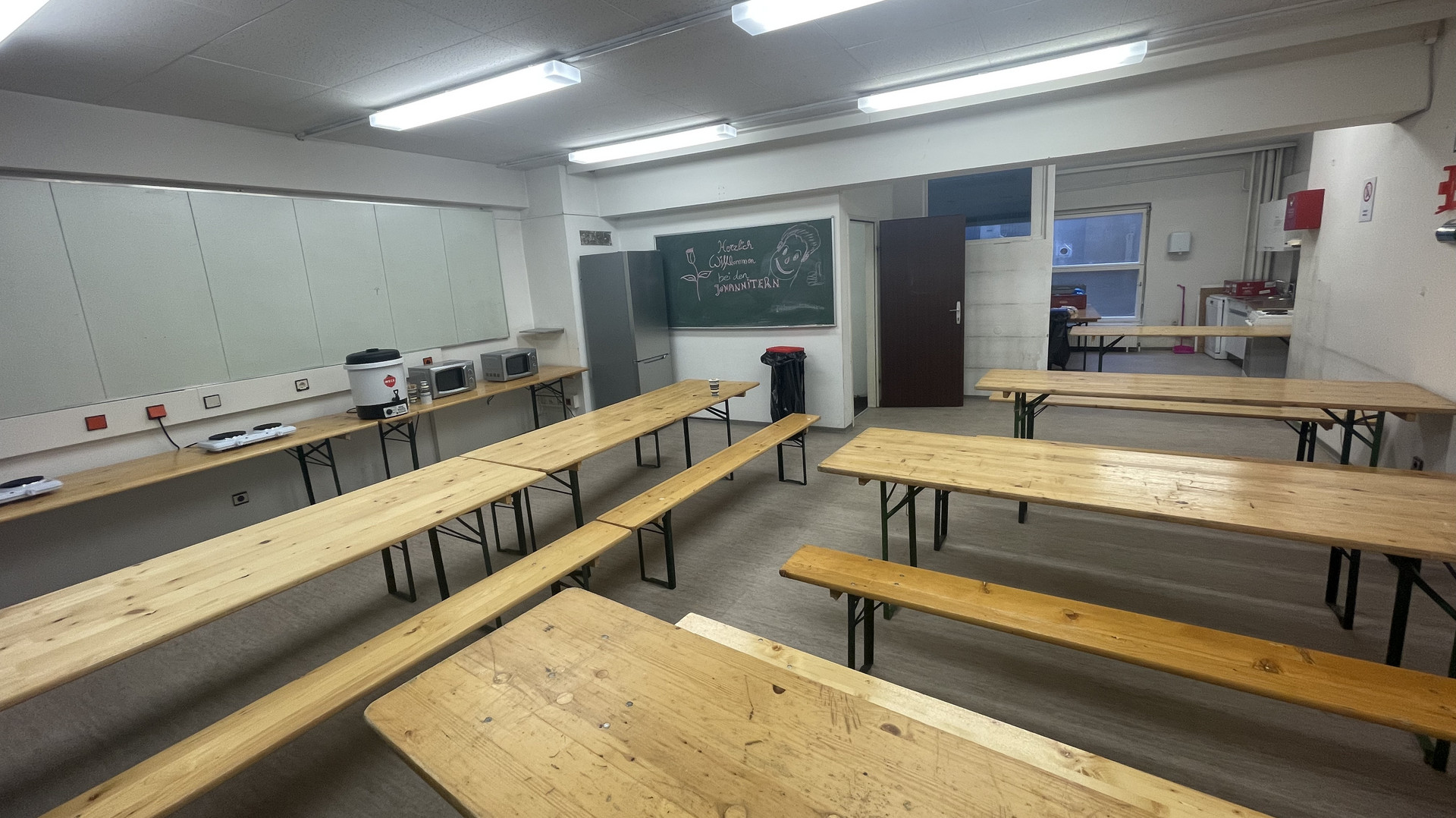 Ein Gemeinschaftsraum mit Heurigengarniture, wo die Menschen essen können. Im Hintergrund stehen Herdplatten, Mikrowellen und ein Kühlschrank.