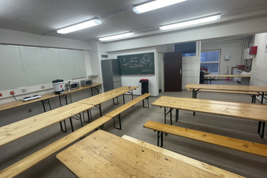 Ein Gemeinschaftsraum mit Heurigengarniture, wo die Menschen essen können. Im Hintergrund stehen Herdplatten, Mikrowellen und ein Kühlschrank.