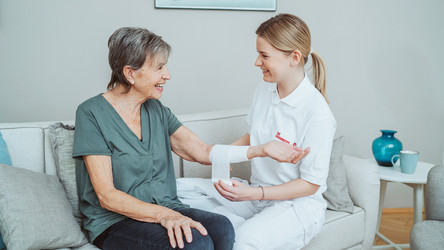 Eine Pflegerin sitzt mit der Patientin auf dem Sofa und legt ihr am Unterarm einen Verband an.