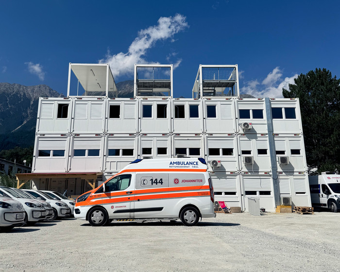 weiße, zweistöckige Container auf Parkplatz. Davor steht ein Johanniter-Rettungsauto.