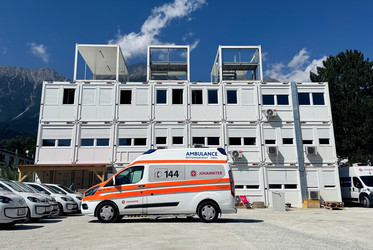 weiße, zweistöckige Container auf Parkplatz. Davor steht ein Johanniter-Rettungsauto.