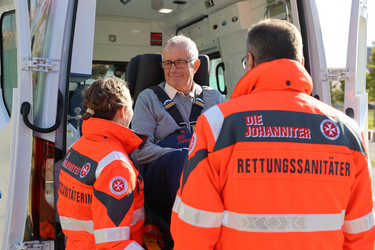 Zwei Johanniter heben einen Patienten auf einer Fahrtrage aus dem Krankenwagen.