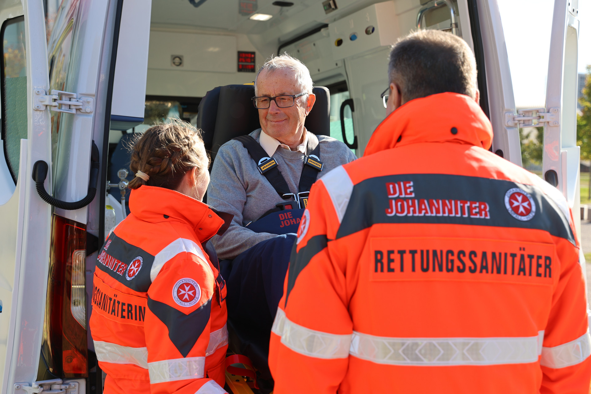 Zwei Johanniter heben einen Patienten auf einer Fahrtrage aus dem Krankenwagen.