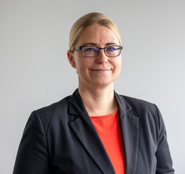 blonde Frau mit Brille steht vor weißer Wand, trägt schwarzes Sakko mit rotem T-Shirt