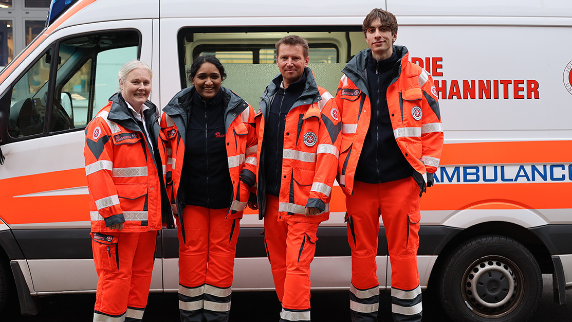 Myuri steht gemeinsam mit dem Team vom Pflegenotdienst vor einem Rettungsauto. 