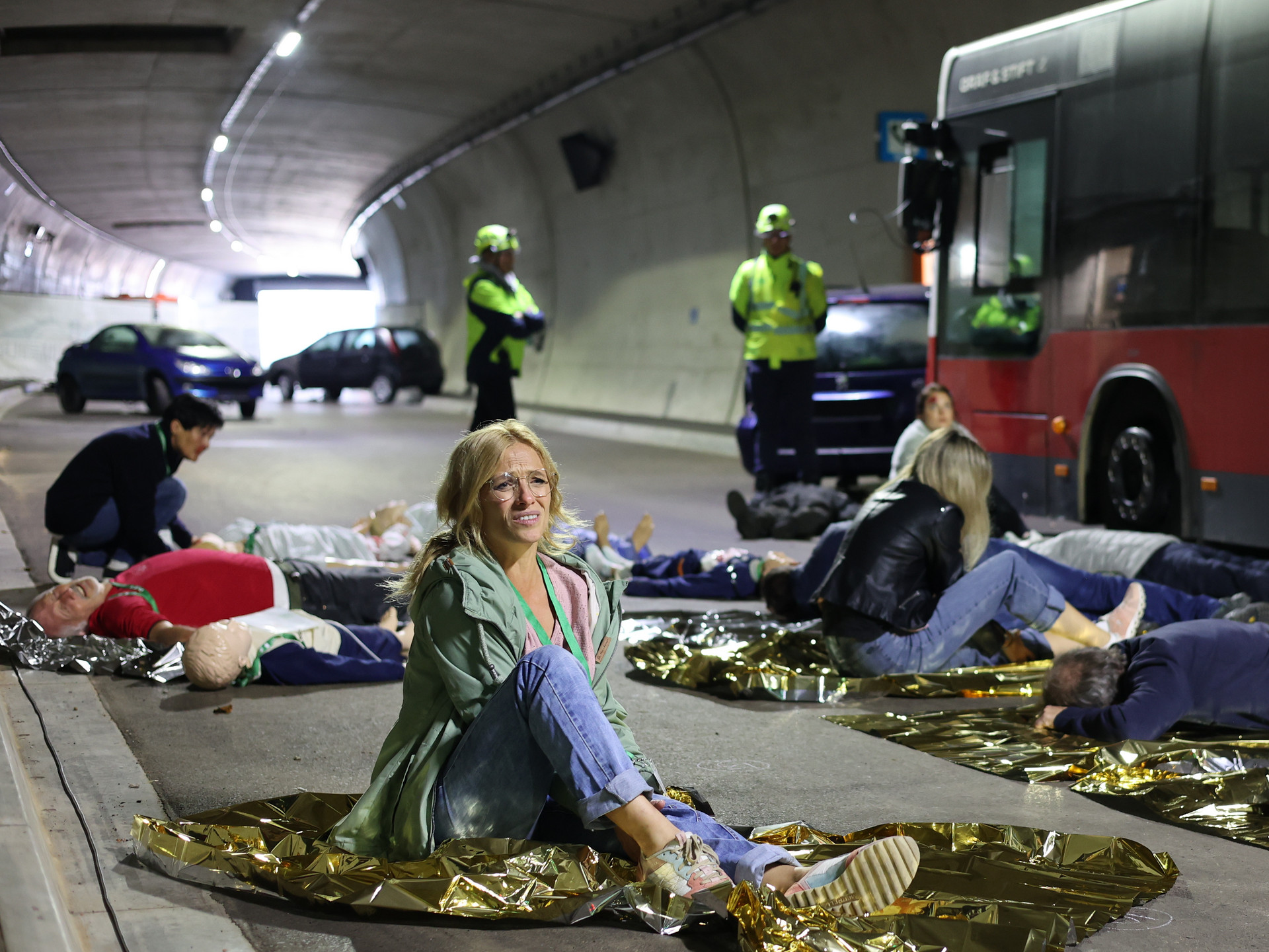 Mehrere Statist:innen mit geschminkten Verletzungen sitzen oder liegen am Boden vor dem verunfallten Bus