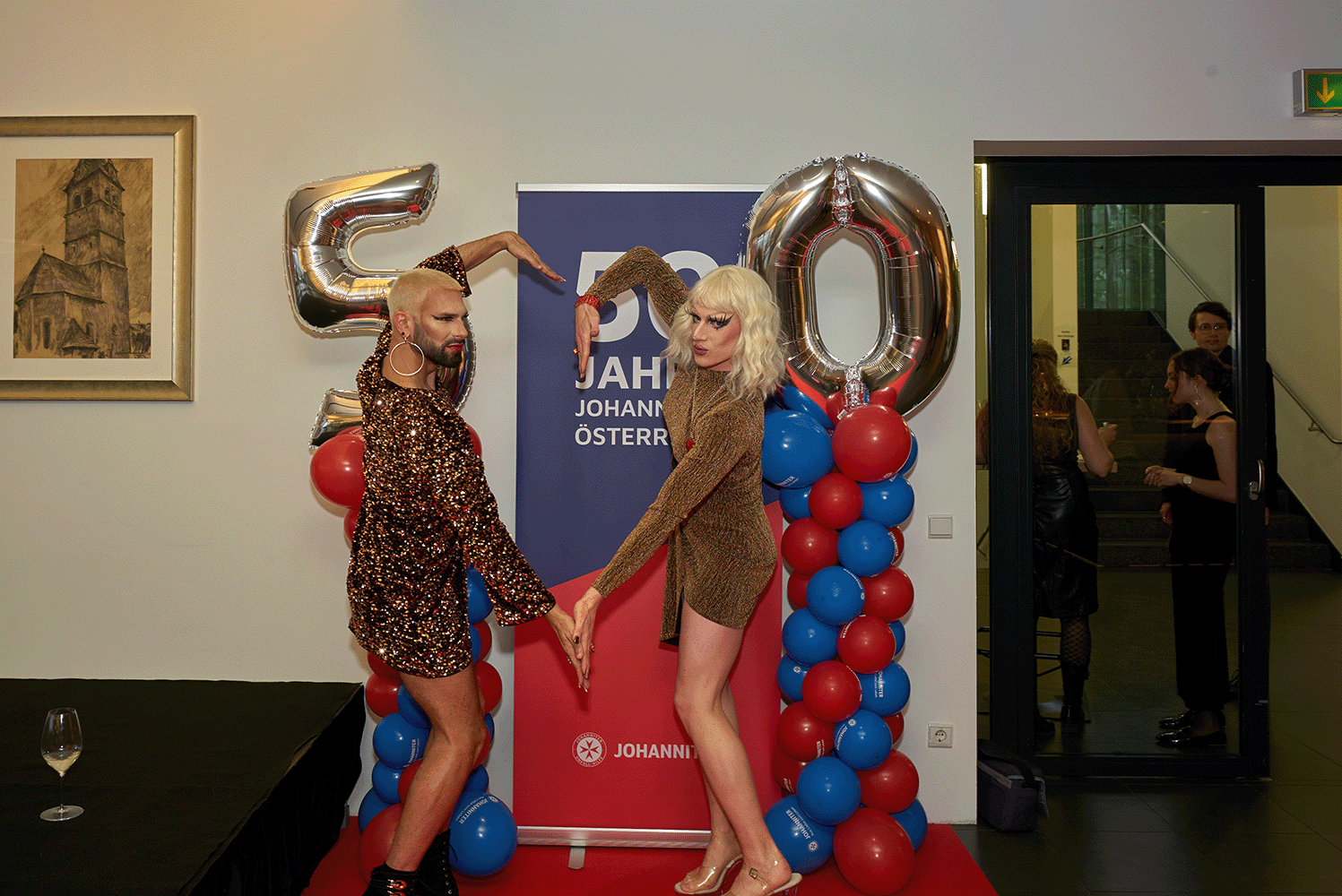 2 Dragqueens stehen vor einem 50er Ballon und machen ein Herz vor der Kamera