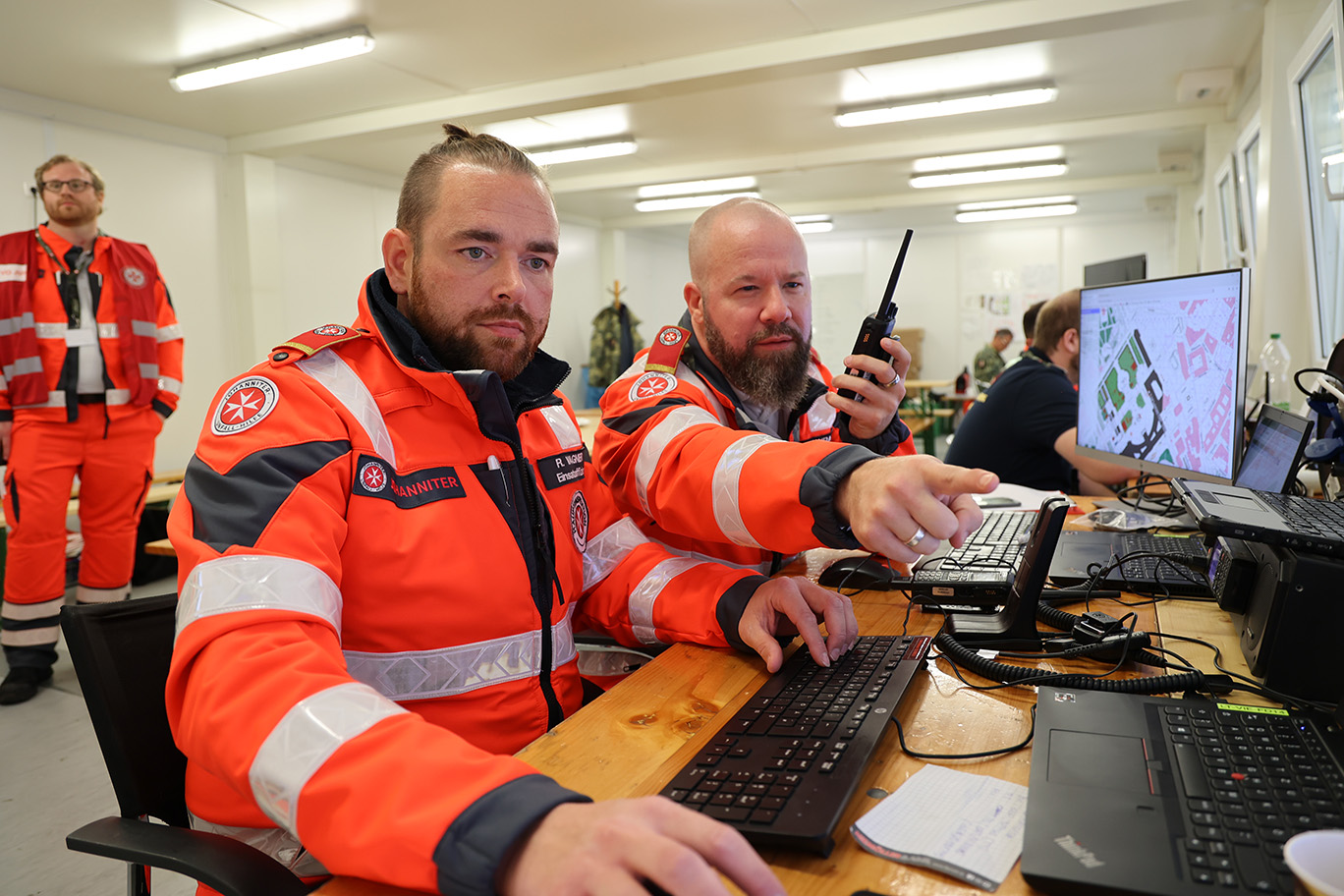 Zwei Johanniter haben am Tisch vor sich Bildschirme, wo die Lage überwacht wird.